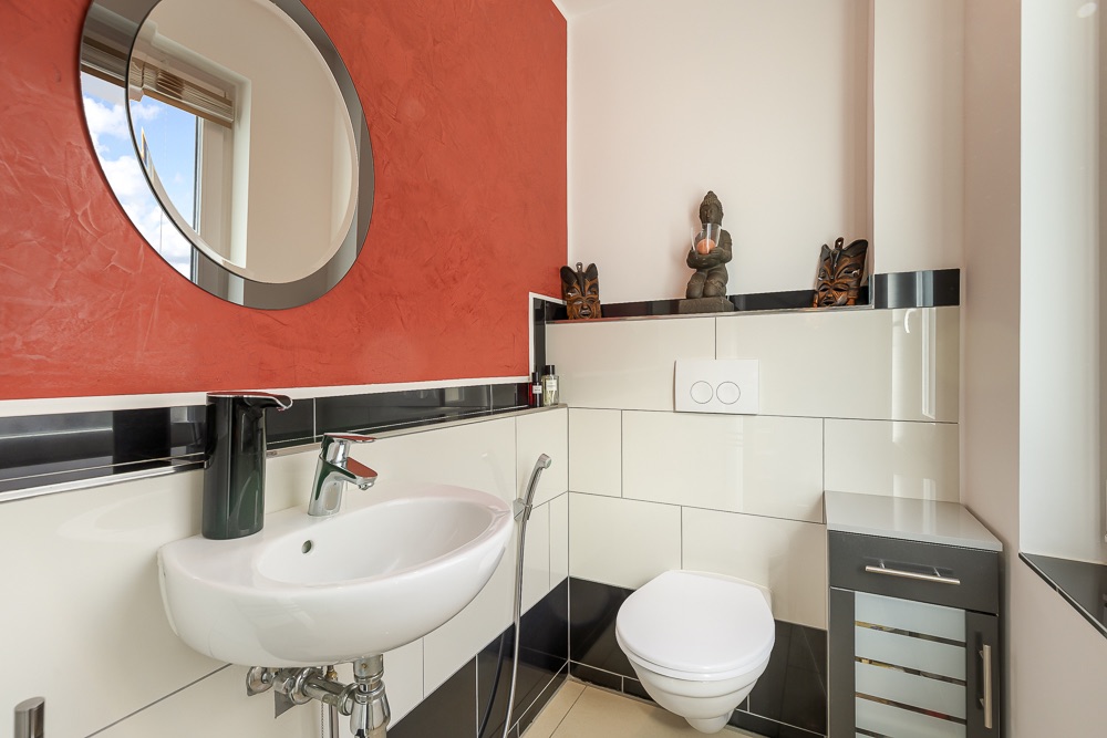 Modernes Badezimmer mit rundem Spiegel, Waschbecken und Wandtoilette, rote Wand und Dekorationen.