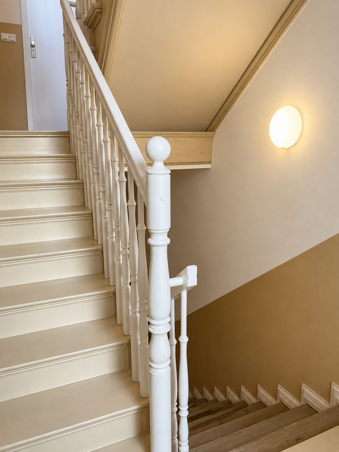 Helle Treppe mit weißem Geländer, Wandlampe und Tür im Hintergrund.