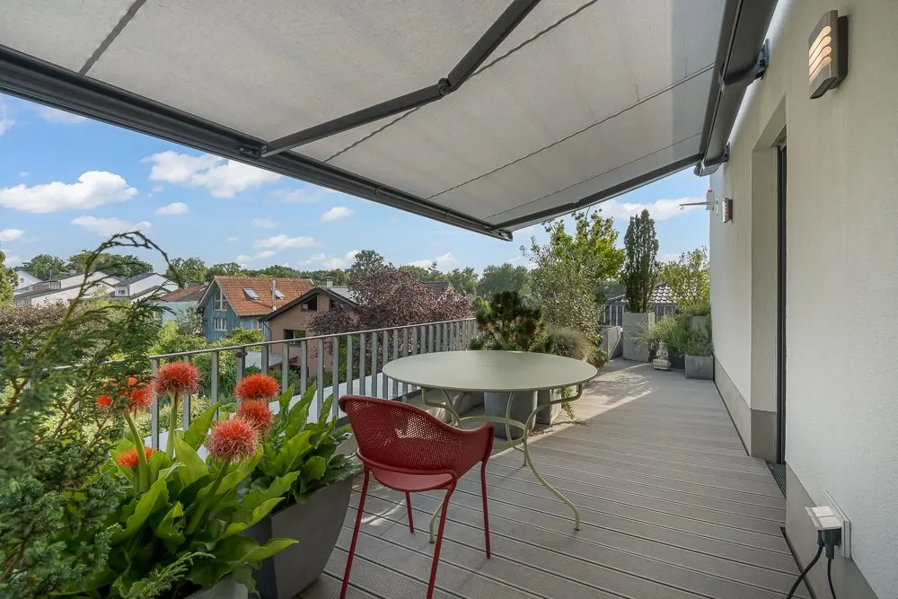 Balkon mit Markise, rundem Tisch, rotem Stuhl und vielen Pflanzen, Ausblick auf Nachbarhäuser.