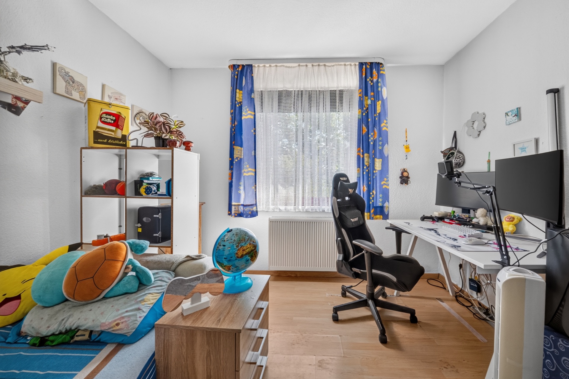 Kinderzimmer mit Bett, Schreibtisch, Stuhl, Regalen und einem Fenster mit blauen Vorhängen.