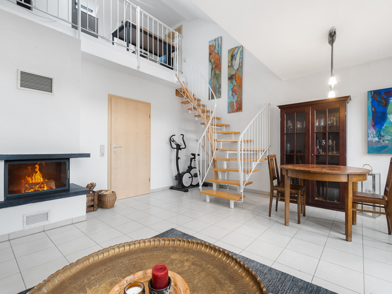 Wohnzimmer mit Kamin, Esstisch, Holztreppe, Vitrine und Trainingsgerät auf weißem Fliesenboden.
