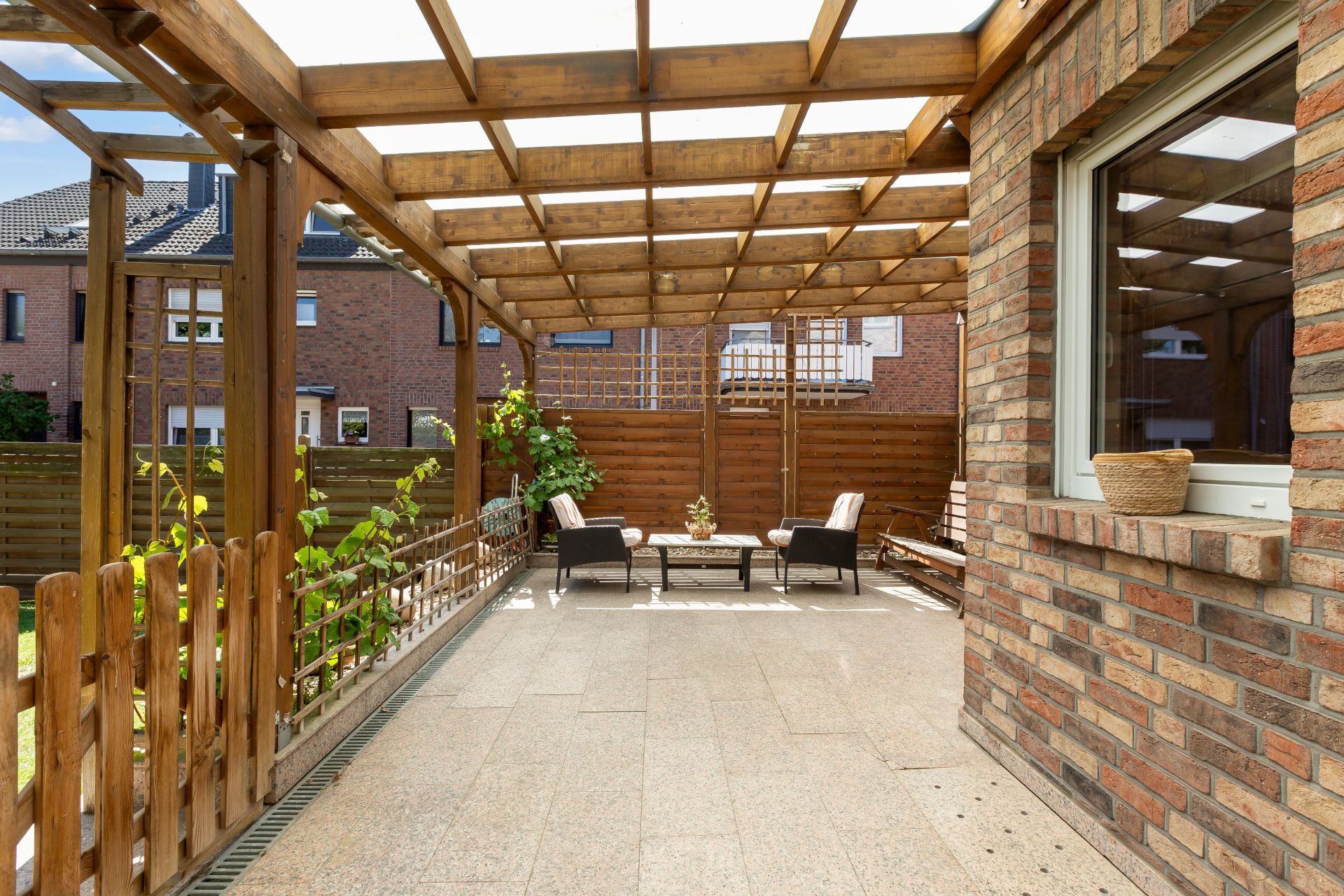 Überdachte Terrasse mit Holzdach, Gartenmöbeln und Pflanzen an einem Backsteinhaus.