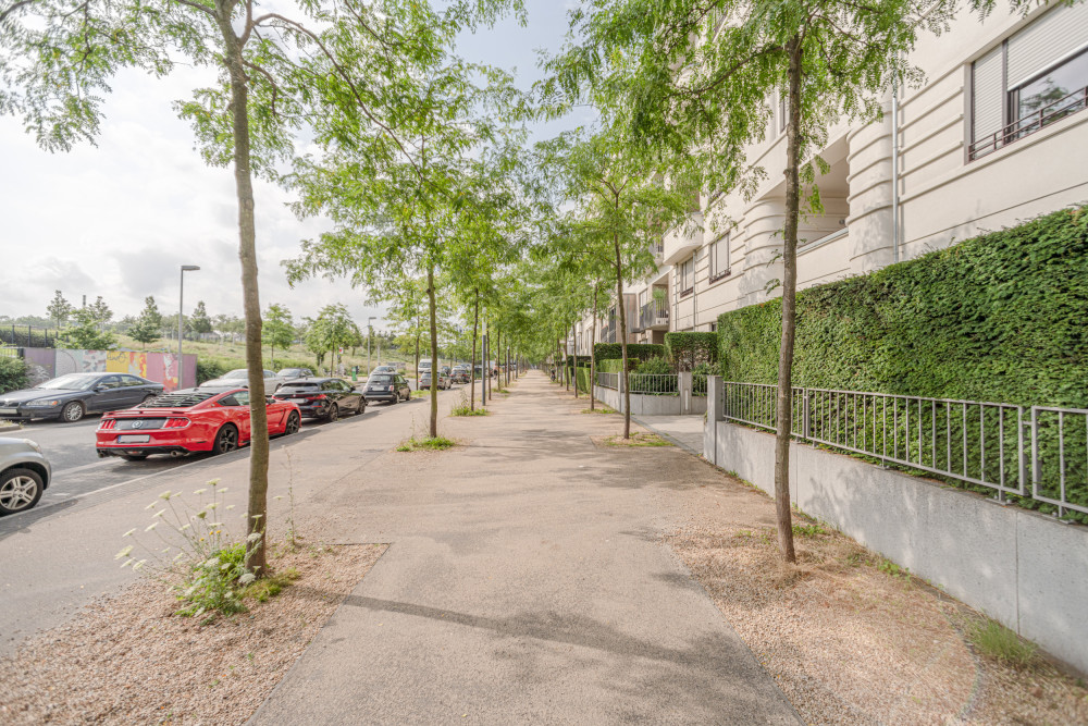 Gehweg mit Bäumen an einer Wohnstraße, Autos parken seitlich, Wohngebäude rechts.