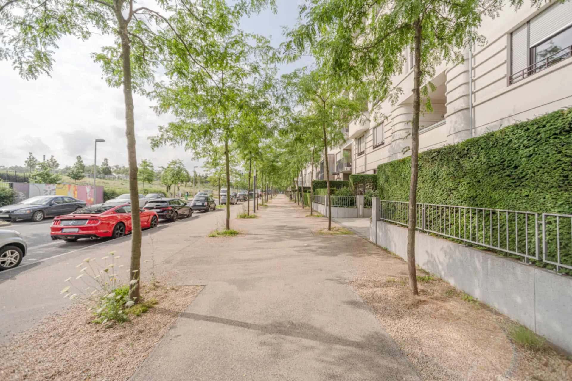 Gehweg mit Bäumen an einer Wohnstraße, Autos parken seitlich, Wohngebäude rechts.