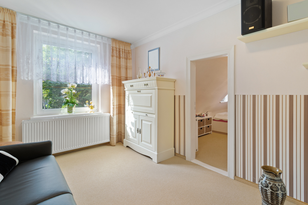 Helles Wohnzimmer mit Sofa, Fenster, Kommode und Tür zu einem weiteren Raum mit Bett.