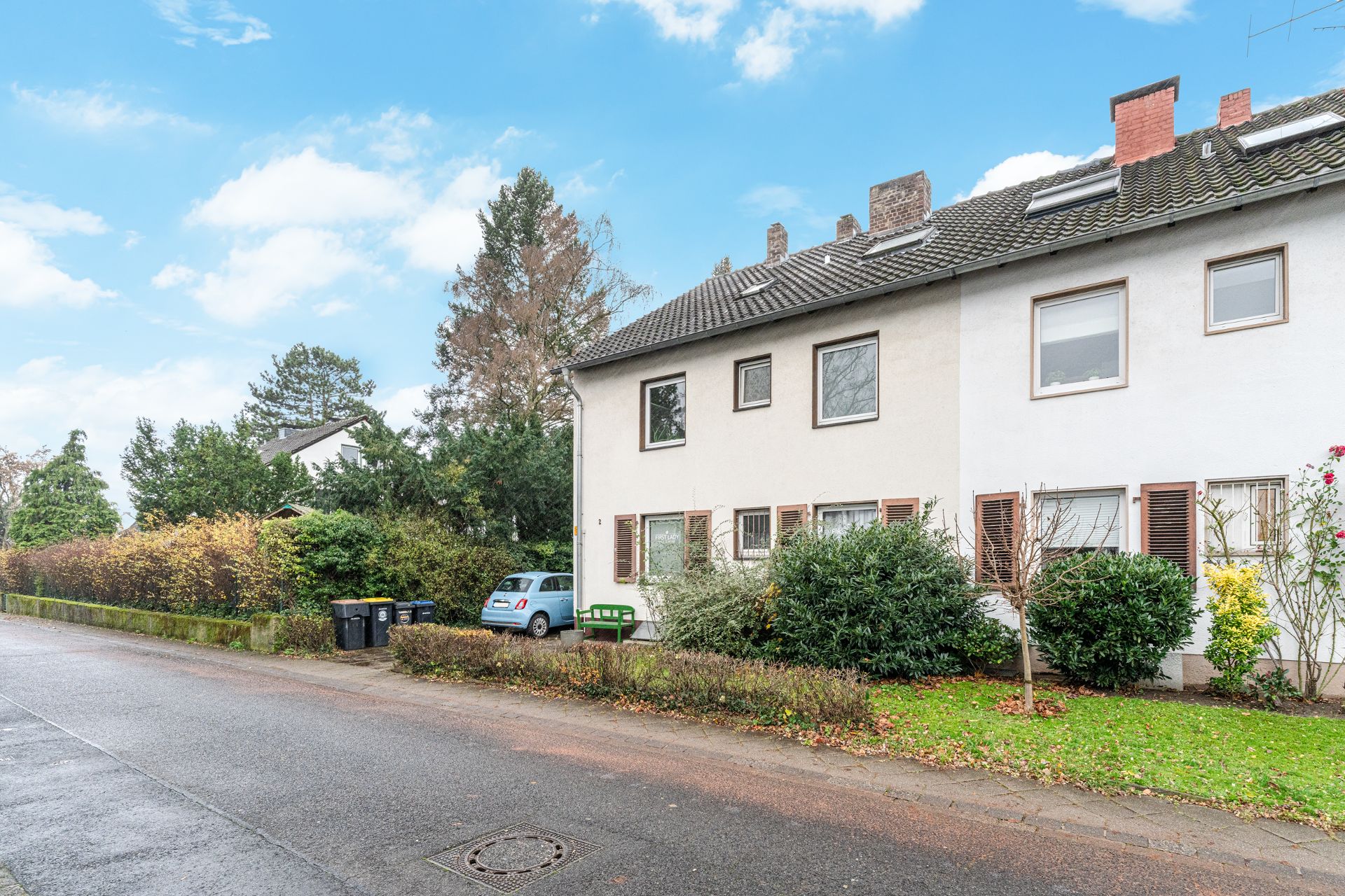 Mehrfamilienhaus mit Vorgarten, parkendem Auto und Mülltonnen an einer ruhigen Straße.
