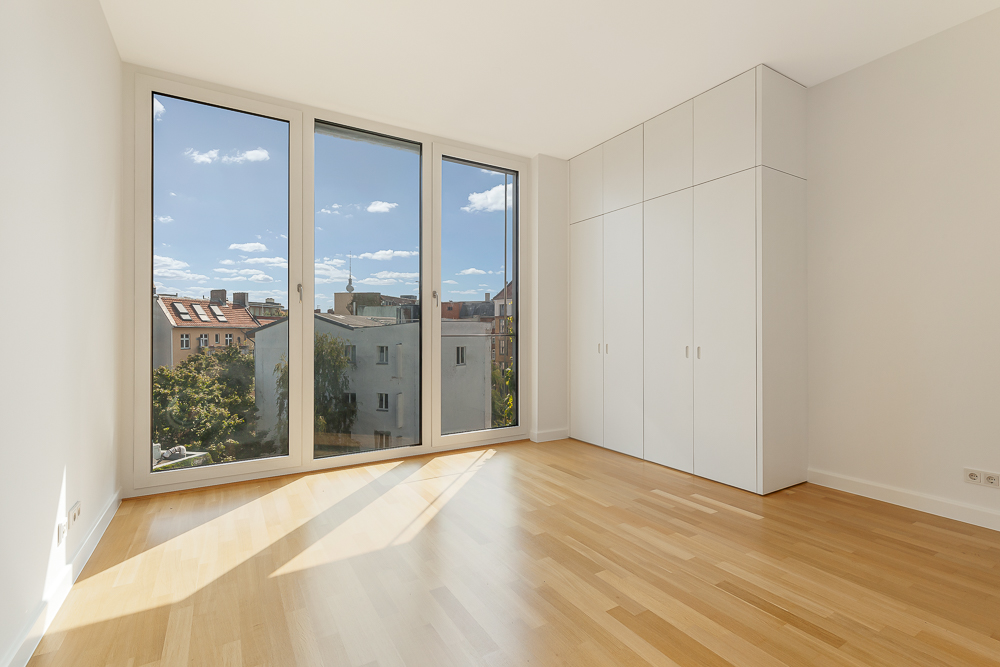 Helles Zimmer mit Holzboden, großen Fenstern und weißem Einbauschrank.