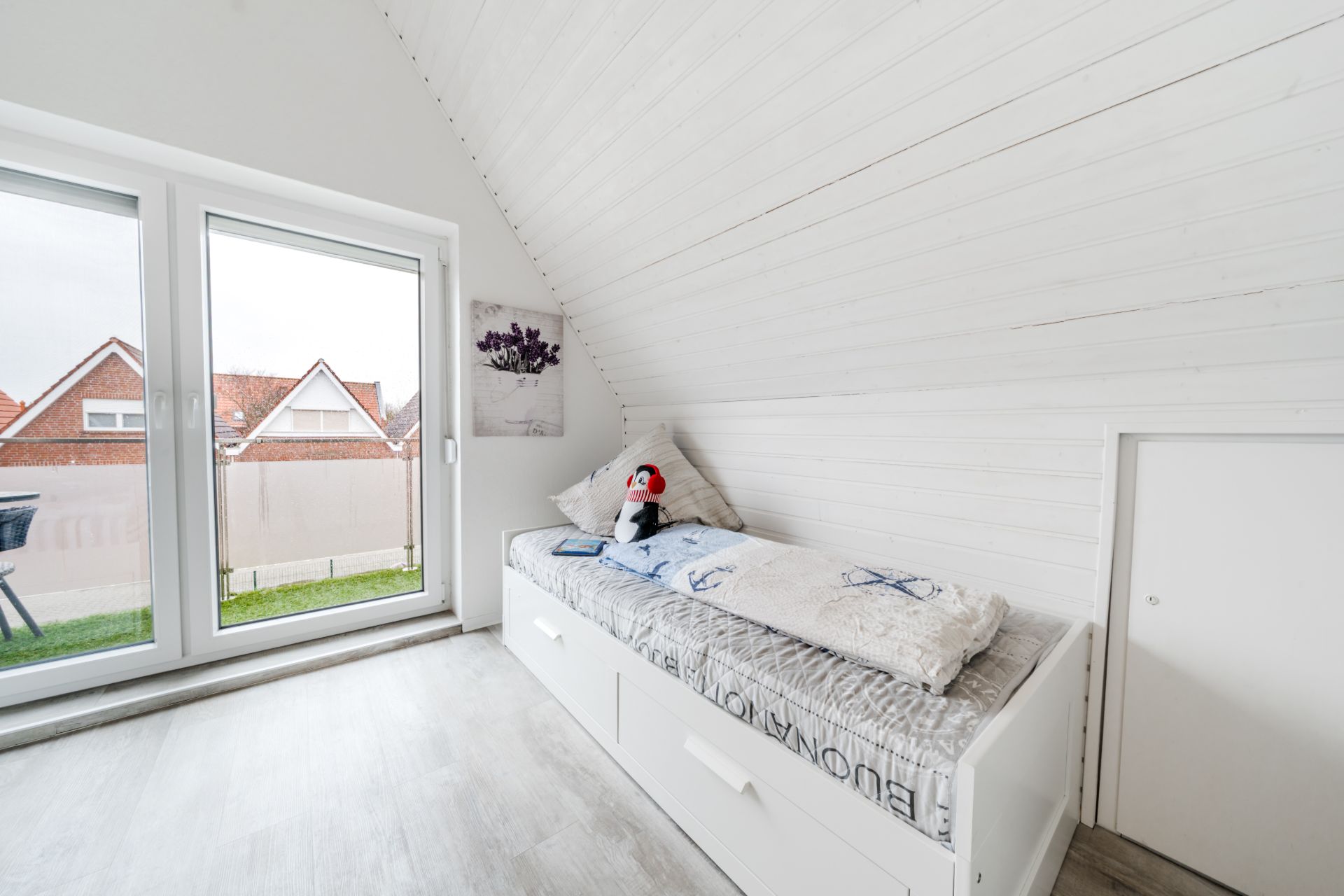 Helles Zimmer mit Einzelbett, großer Fensterfront und weißer Holzvertäfelung.