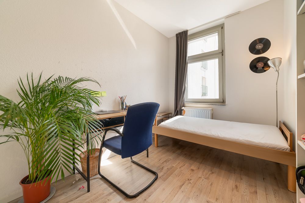 Helles Zimmer mit Einzelbett, Schreibtisch, blauem Stuhl, Flur, Pflanze und Fenster.