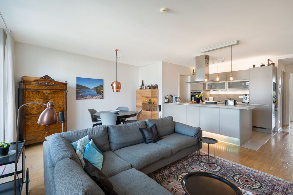 Helles Wohnzimmer mit grauem Ecksofa, Teppich, Esstisch und offener moderner Küche.