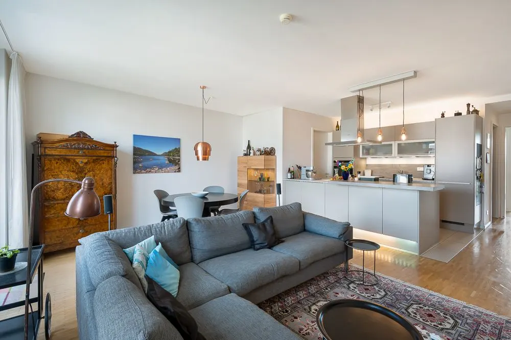 Helles Wohnzimmer mit grauem Ecksofa, Teppich, Esstisch und offener moderner Küche.