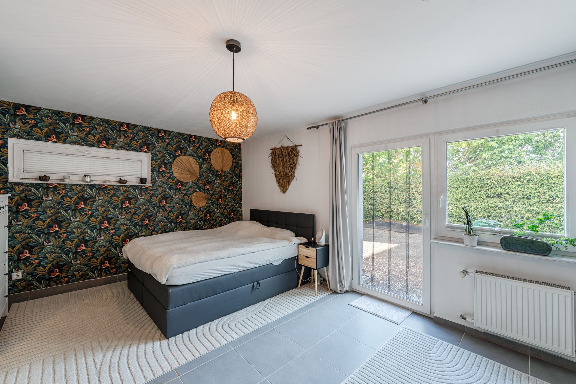 Schlafzimmer mit gemusteter Tapete, Doppelbett, Nachttisch und großen Fenstern zum Garten.