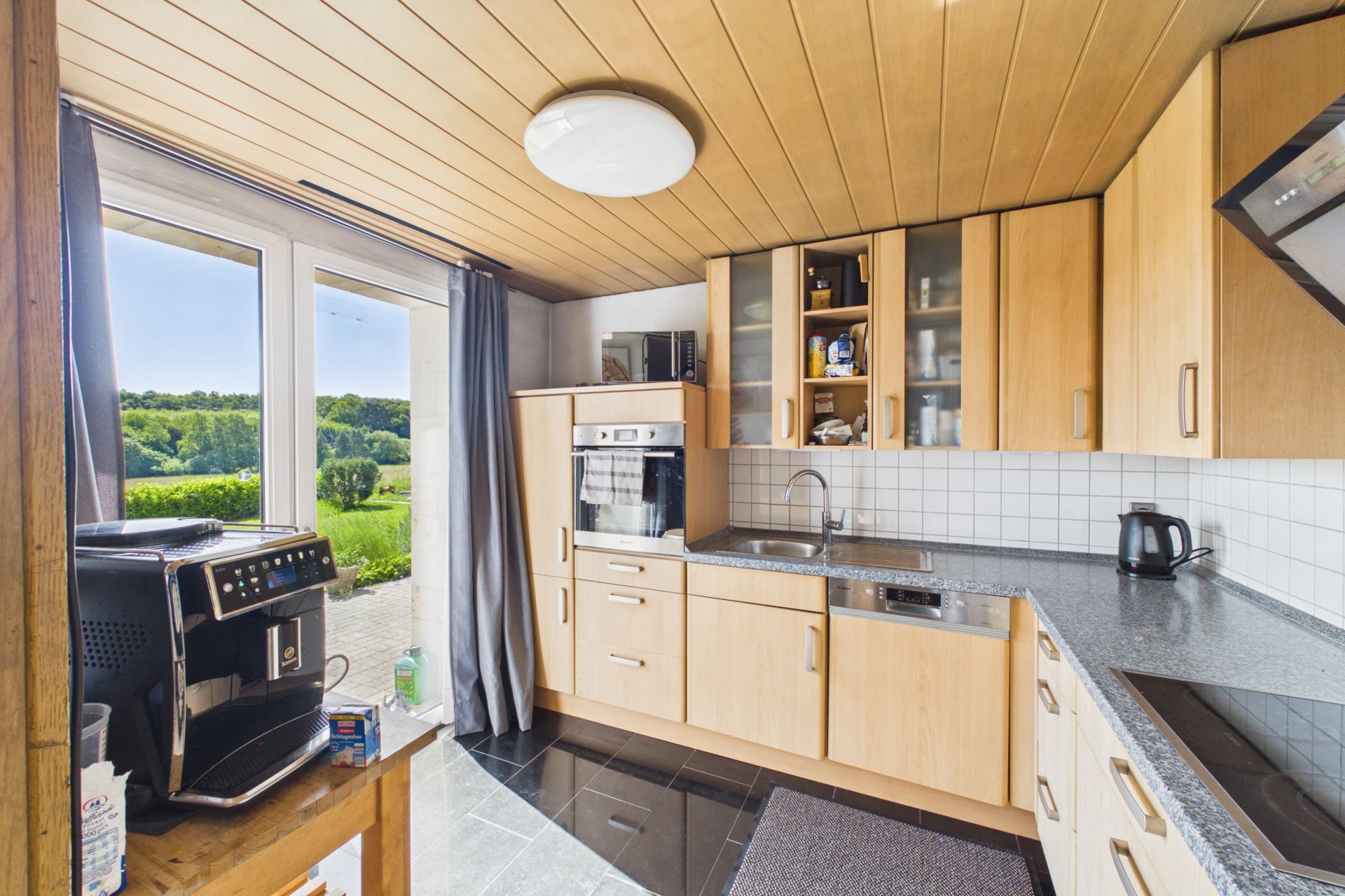 Moderne Küche mit Holzschränken, großem Fenster, Kaffeemaschine und Blick ins Grüne.