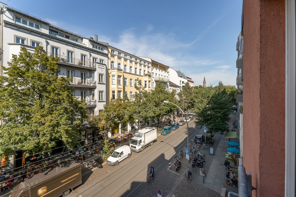 Blick auf eine belebte Straße mit Wohnhäusern, Bäumen, Autos und Fahrrädern bei sonnigem Wetter.