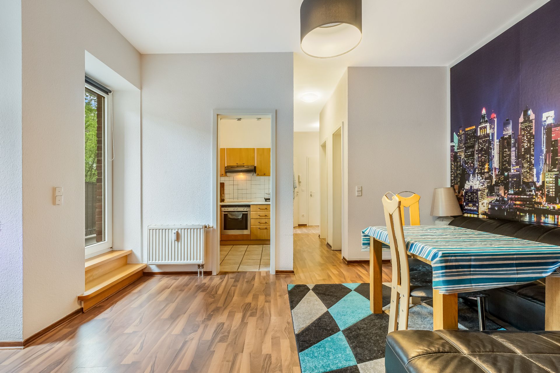 Wohnzimmer mit Esstisch, gemustertem Teppich, Tür zur Küche und Wandbild einer Skyline.