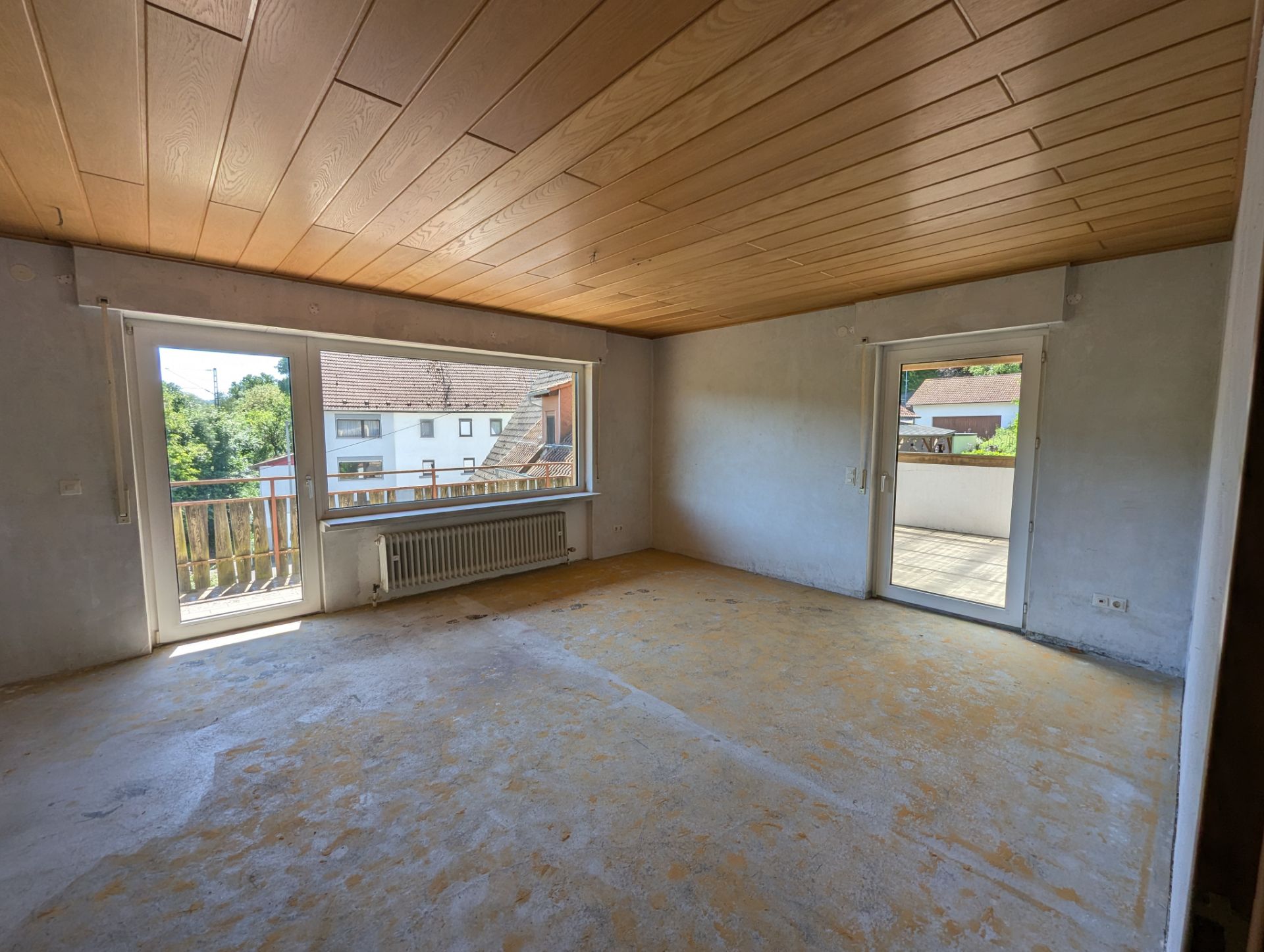 Leeres Zimmer mit Holzdecke, großen Fenstern und Zugang zu einem Balkon.