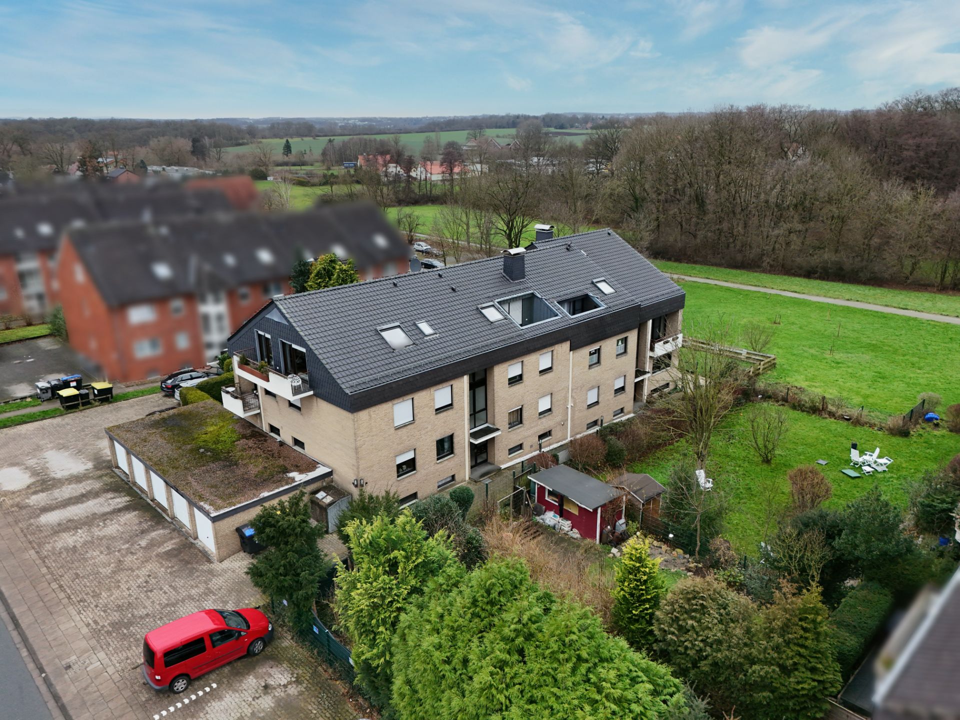 Mehrfamilienhaus mit Garten und Garagen, daneben ein rotes Auto und viel Grün im Hintergrund.