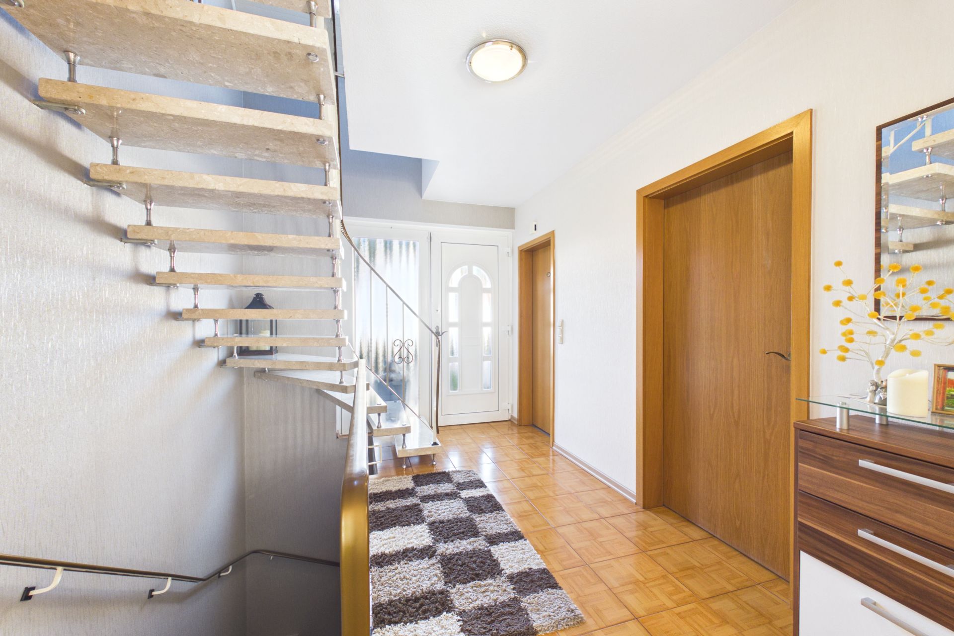 Heller Flur mit offener Treppe, Teppich und Holztüren, Kommode rechts, Haustür im Hintergrund.