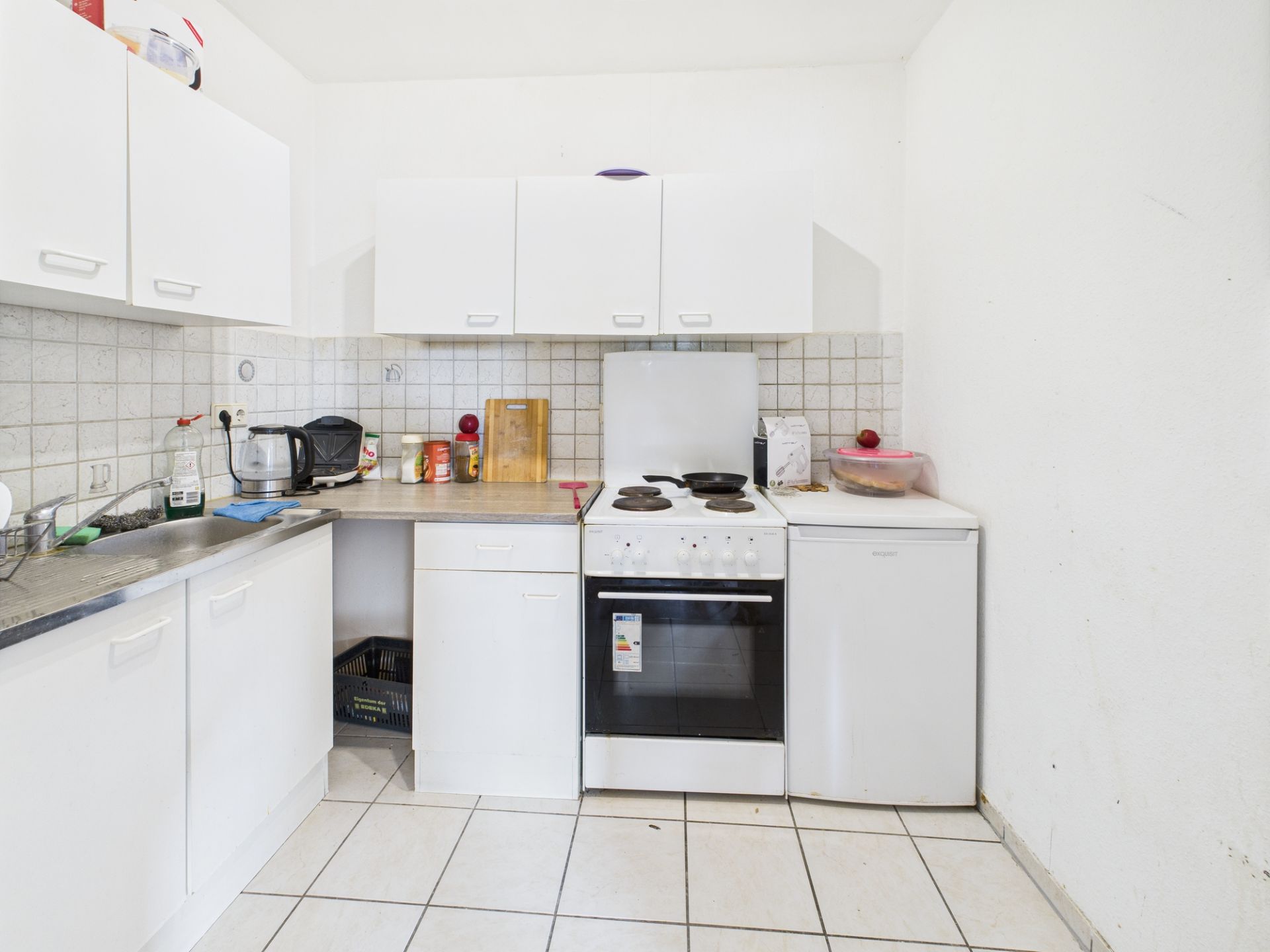 Weiße Einbauküche mit Herd, Spüle, Kühlschrank und Hängeschränken, alles auf weißen Fliesen.