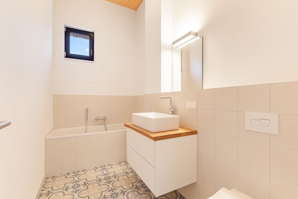 Helles Badezimmer mit Badewanne, Waschbecken, Spiegel und gemustertem Fliesenboden.