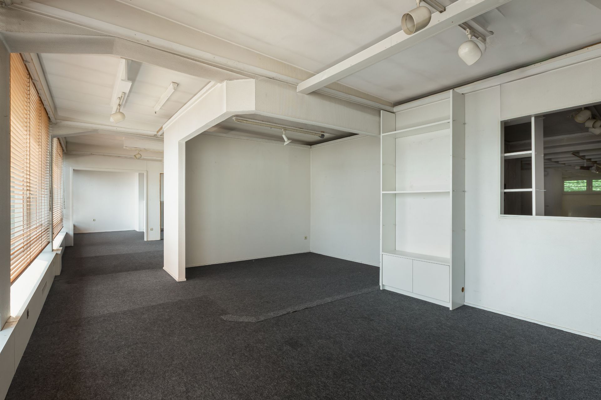 Leerer Raum mit großen Fenstern, grauem Teppich und weißen Wänden, Regal an der rechten Wand.