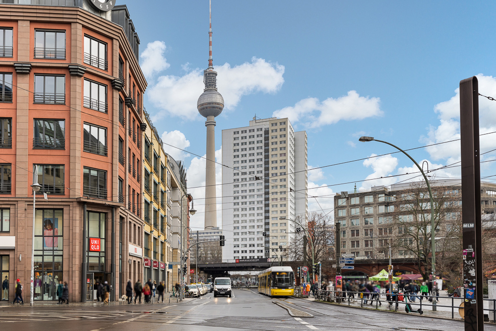 Stadtstraße mit Straßenbahn, Fußgängern und Fernsehturm im Hintergrund bei Tag.