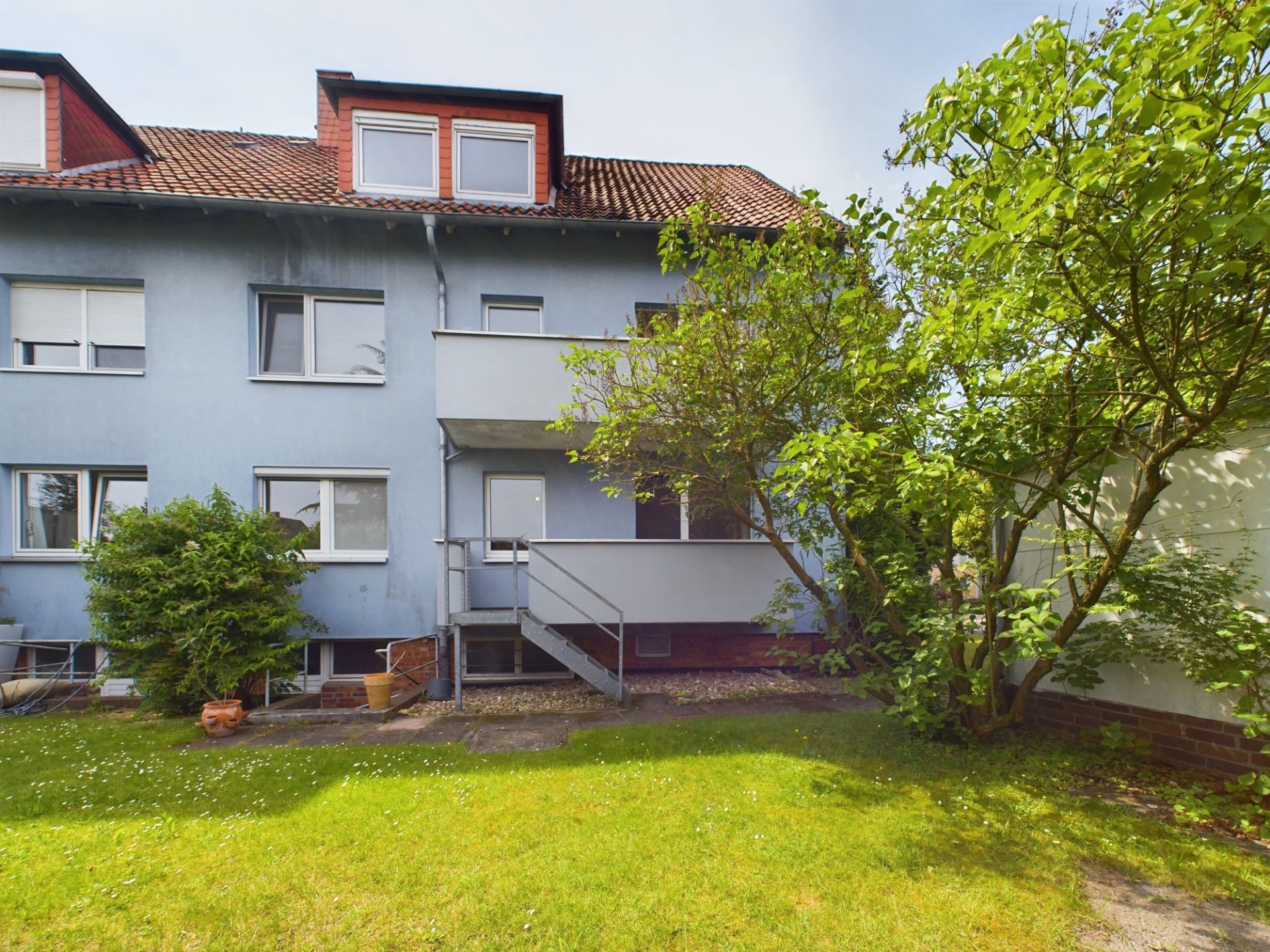 Mehrfamilienhaus mit blauem Putz, Balkon, Gartentreppe und grünem Garten mit Bäumen.