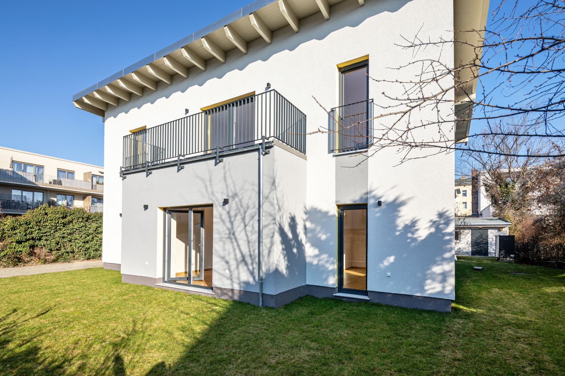 Moderne weiße Wohnhausfassade mit Balkon und großen Fenstern, umgeben von grünem Garten.