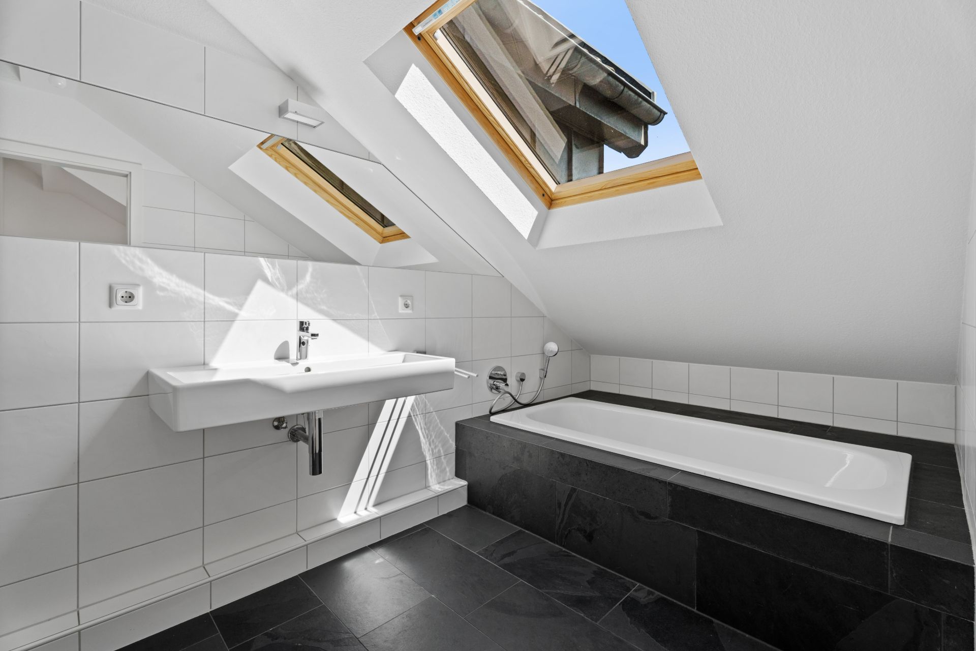 Modernes Badezimmer mit Dachschräge, Badewanne, Waschbecken und zwei Dachfenstern.