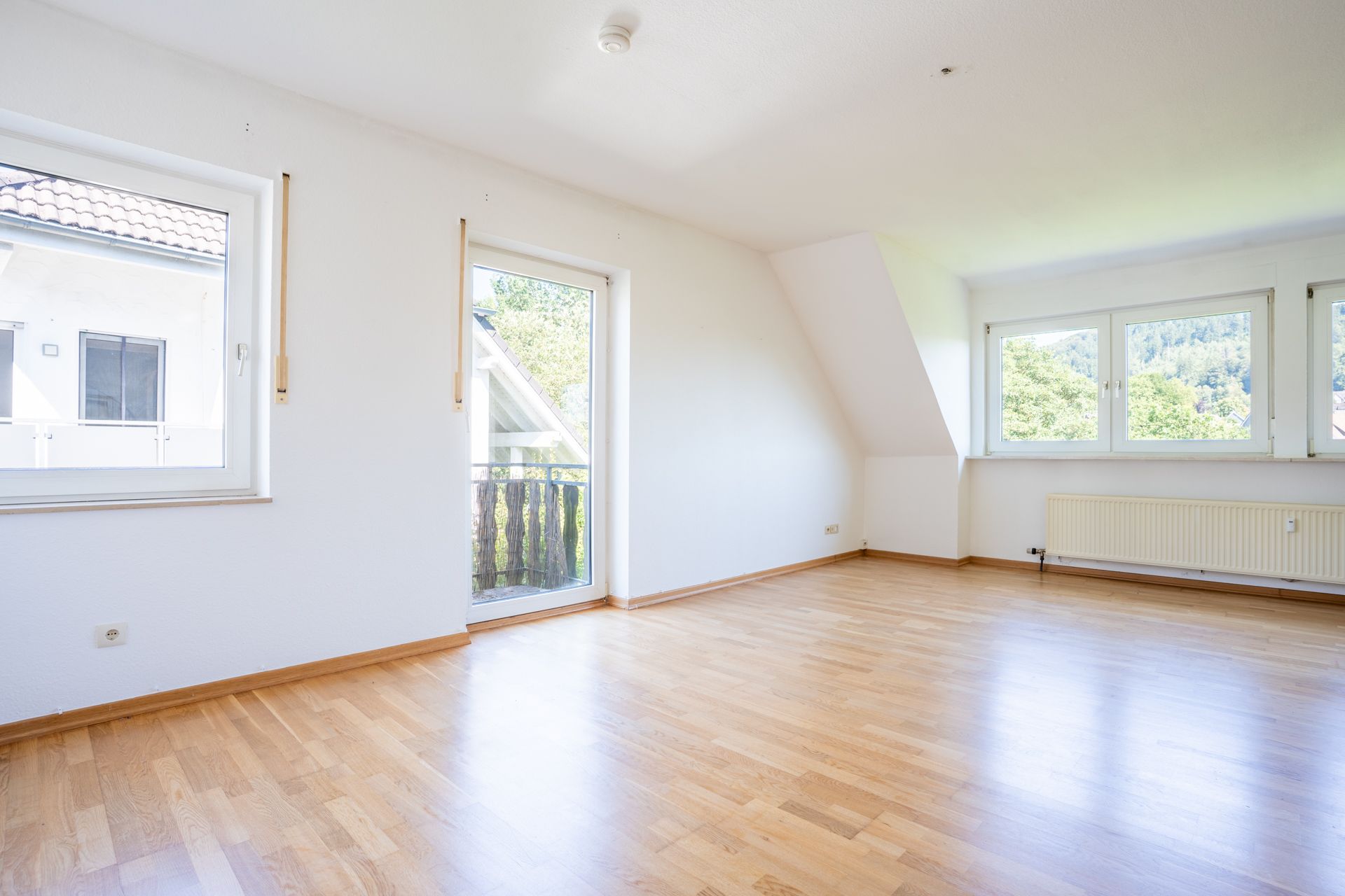Heller leerer Raum mit Parkettboden, großen Fenstern und Tür zum kleinen Balkon.