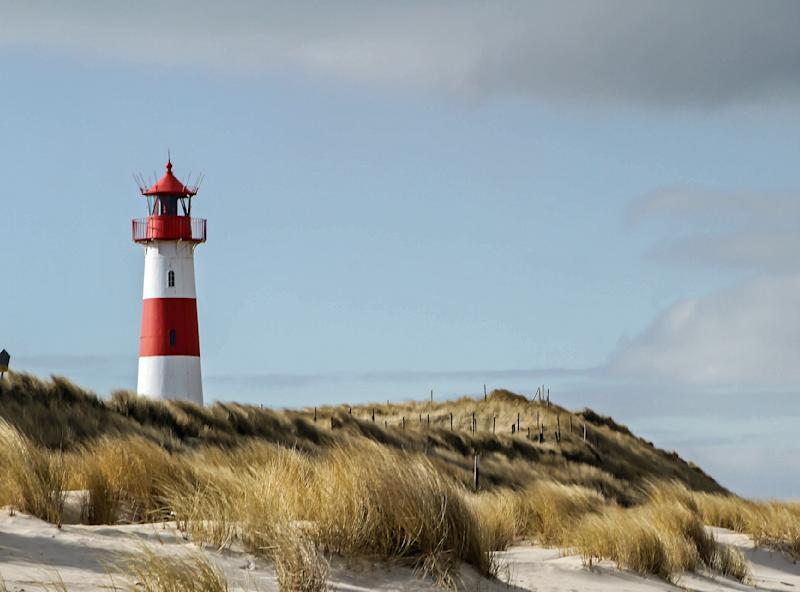 Leuchtturm mit roten und weißen Streifen steht hinter einer Sanddüne mit Gras.