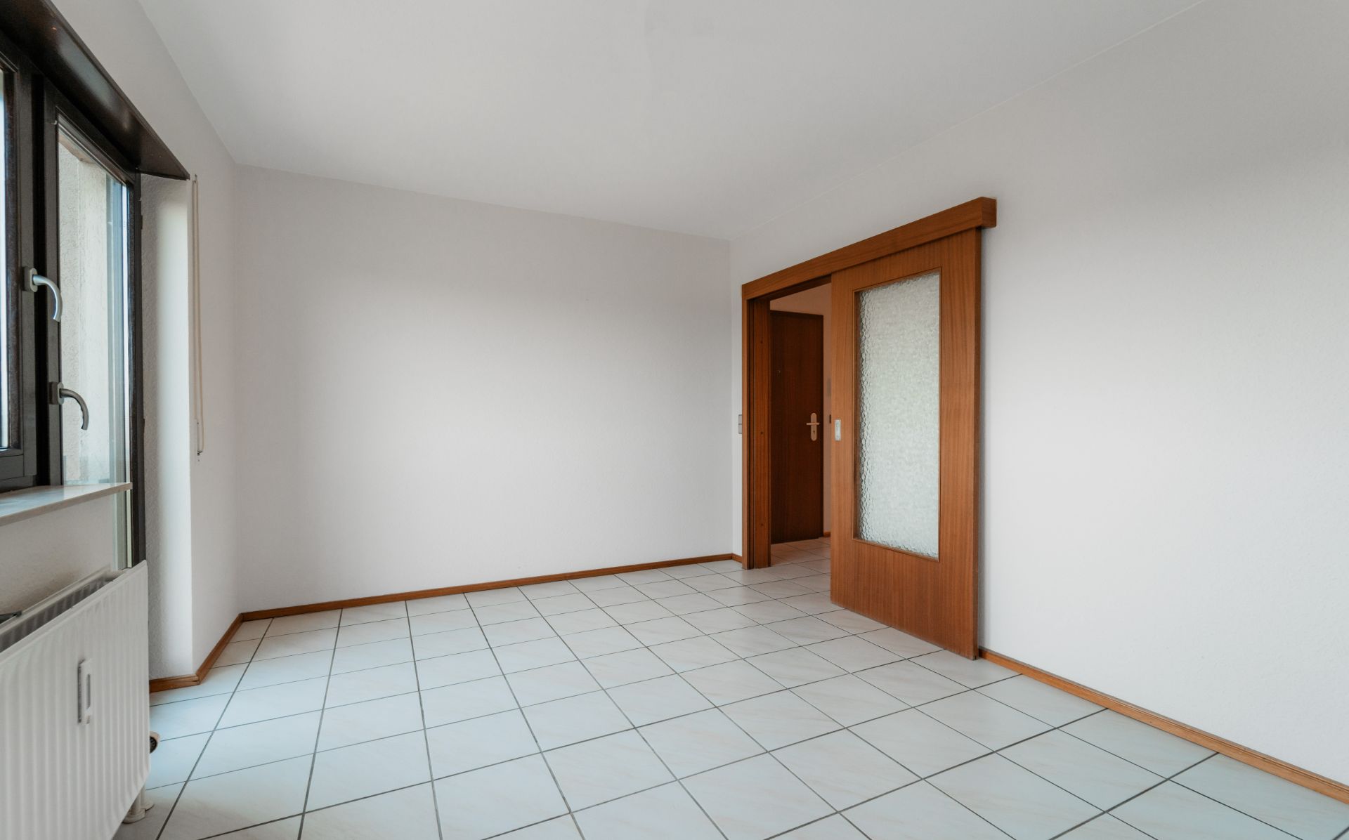 Leerer, heller Raum mit Fliesenboden, großem Fenster und Holzschiebetür.