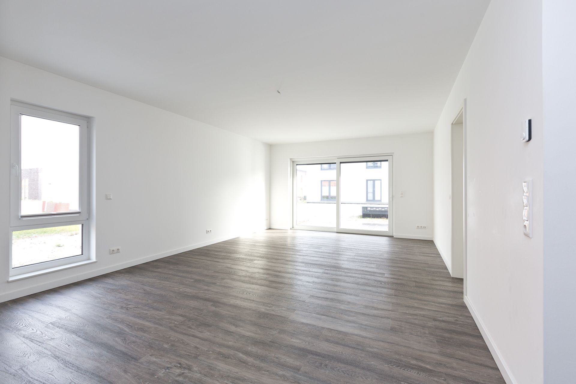 Leeres, helles Wohnzimmer mit großen Fenstern und dunklem Holzboden.