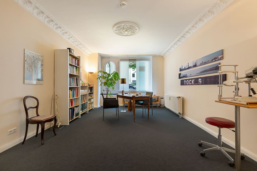 Heller Raum mit Schreibtisch, Bücherregalen, mehreren Stühlen und einem großen Fenster.