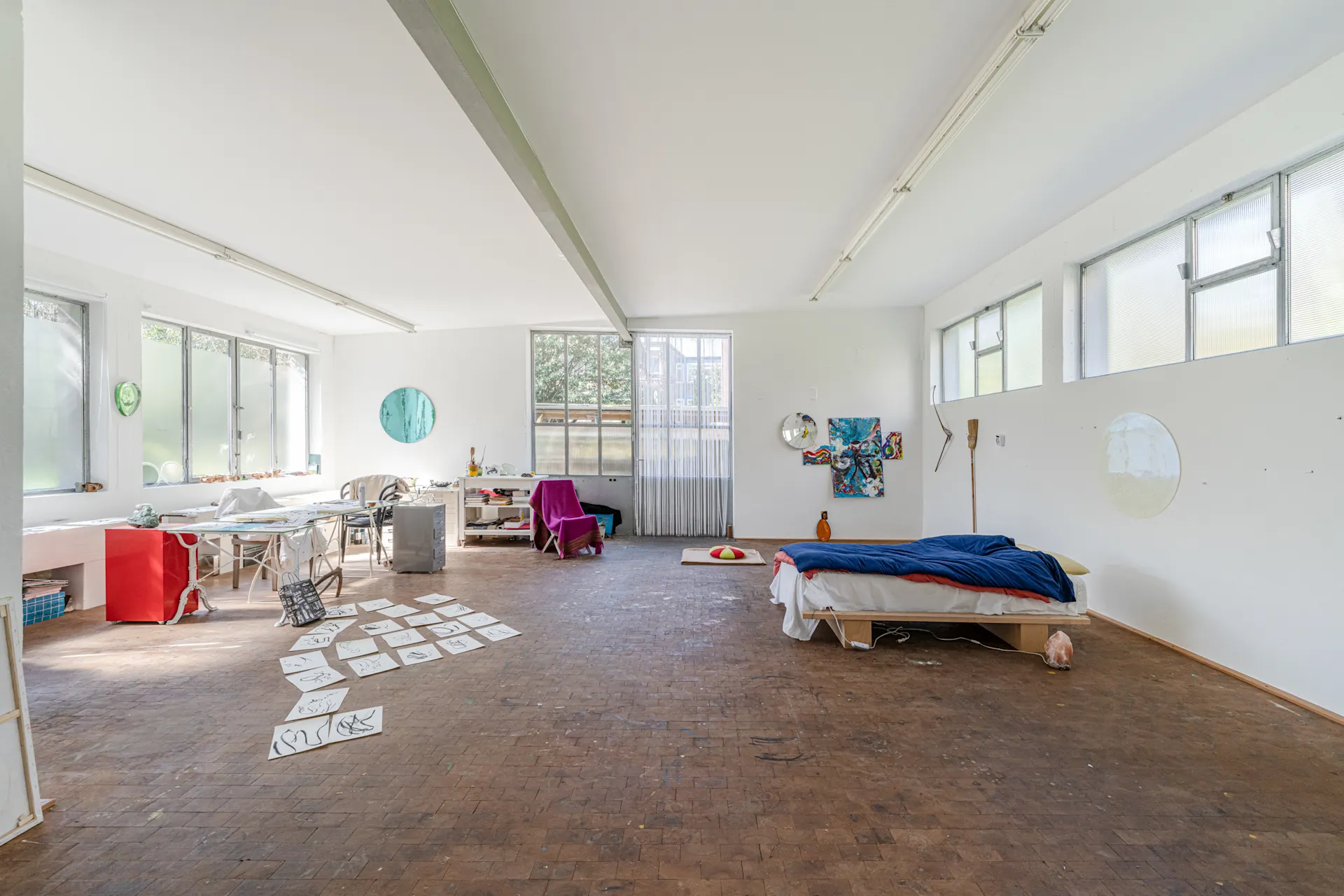 Großer, heller Raum mit Bett, Schreibtisch, Stuhl und Kunstwerken an den Wänden.
