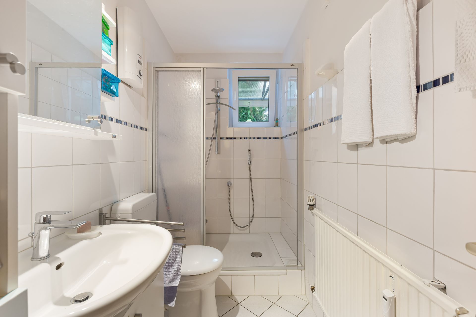 Modernes, weiß gefliestes Badezimmer mit Dusche, Waschbecken, WC und Handtüchern an der Wand.