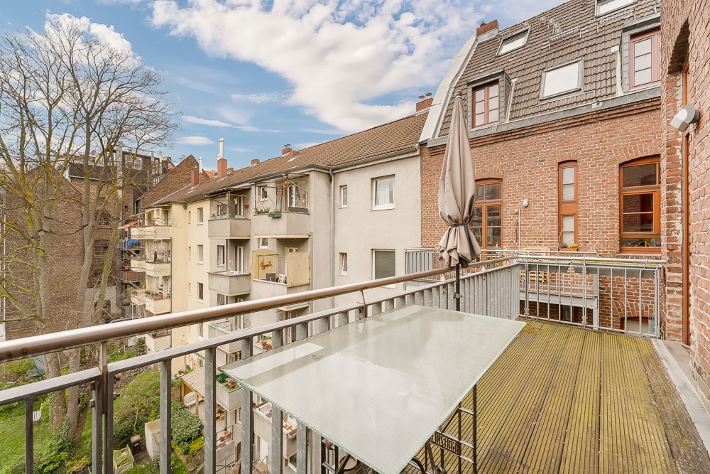 Balkon mit Glastisch und Sonnenschirm, Blick auf Nachbarhäuser und Bäume.