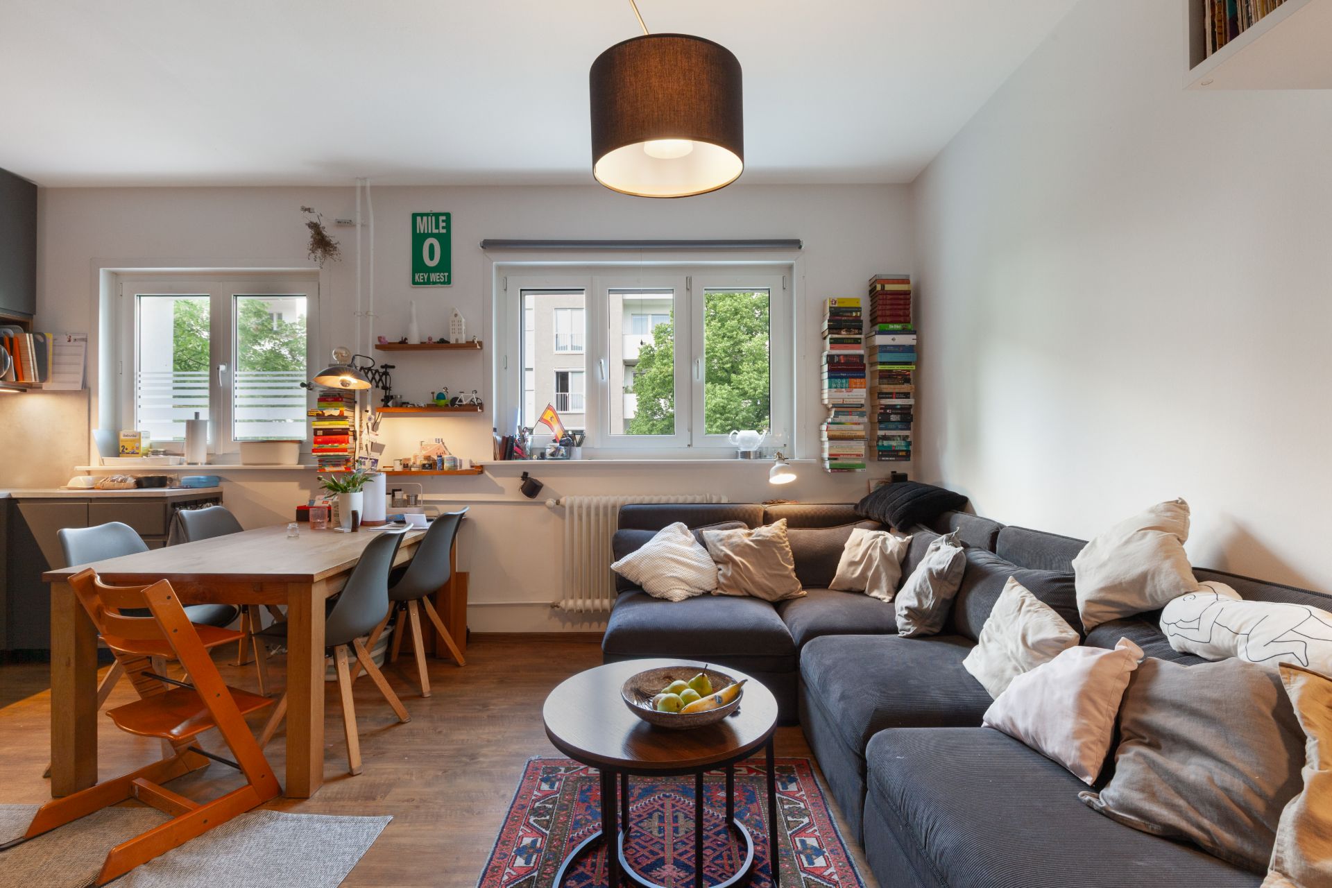 Wohnzimmer mit Ecksofa, Esstisch, Bücherstapeln und großen Fenstern.