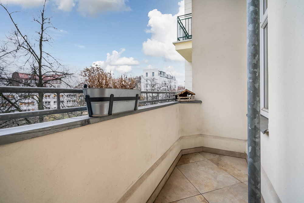 Kleiner Balkon mit Fliesenboden, Geländer, Pflanzkasten und Blick auf Bäume und Gebäude.