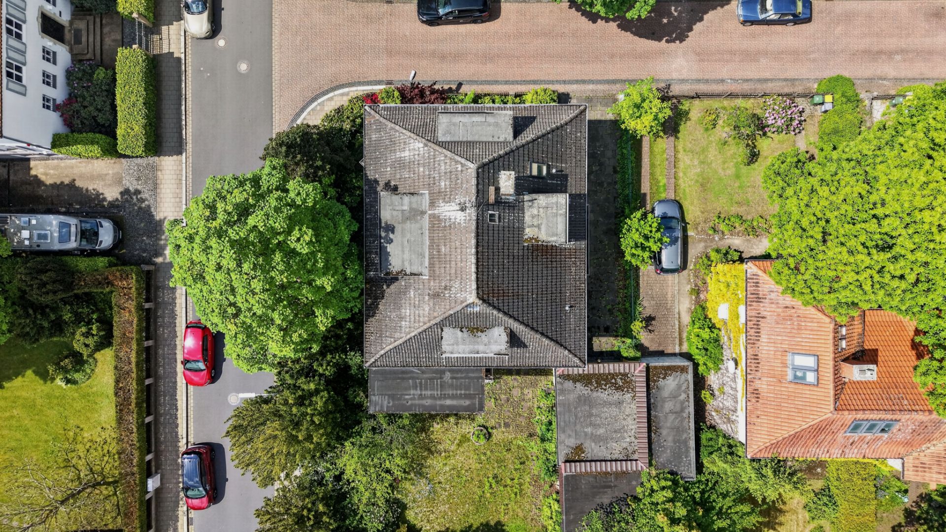 Luftaufnahme eines Wohnhauses mit Garten, umgeben von Bäumen und geparkten Autos auf der Straße.