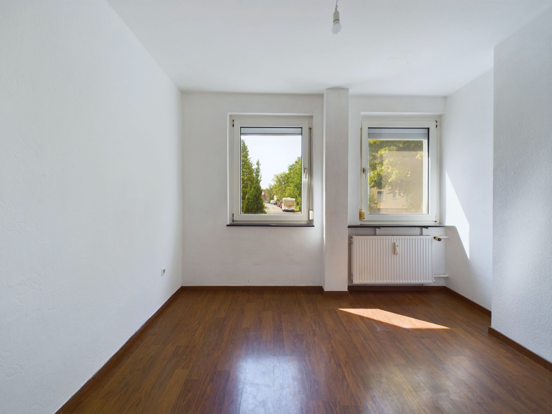 Leerer Raum mit zwei Fenstern, weiß gestrichenen Wänden und braunem Laminatboden.