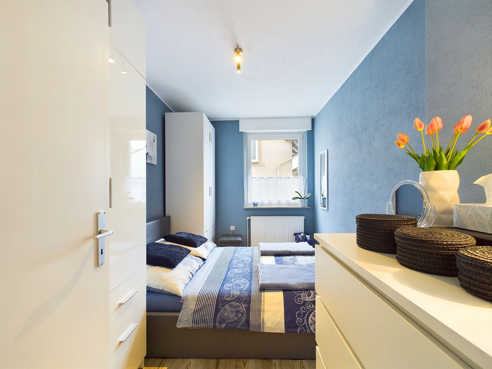 Modernes Schlafzimmer mit blau gestrichenen Wänden, Doppelbett, Kommode und Fenster.