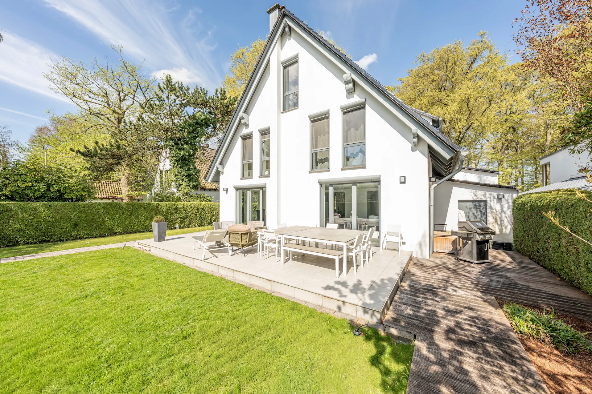 Modernes weißes Haus mit großer Terrasse, Gartenmöbeln und gepflegtem Rasen im Sonnenschein.