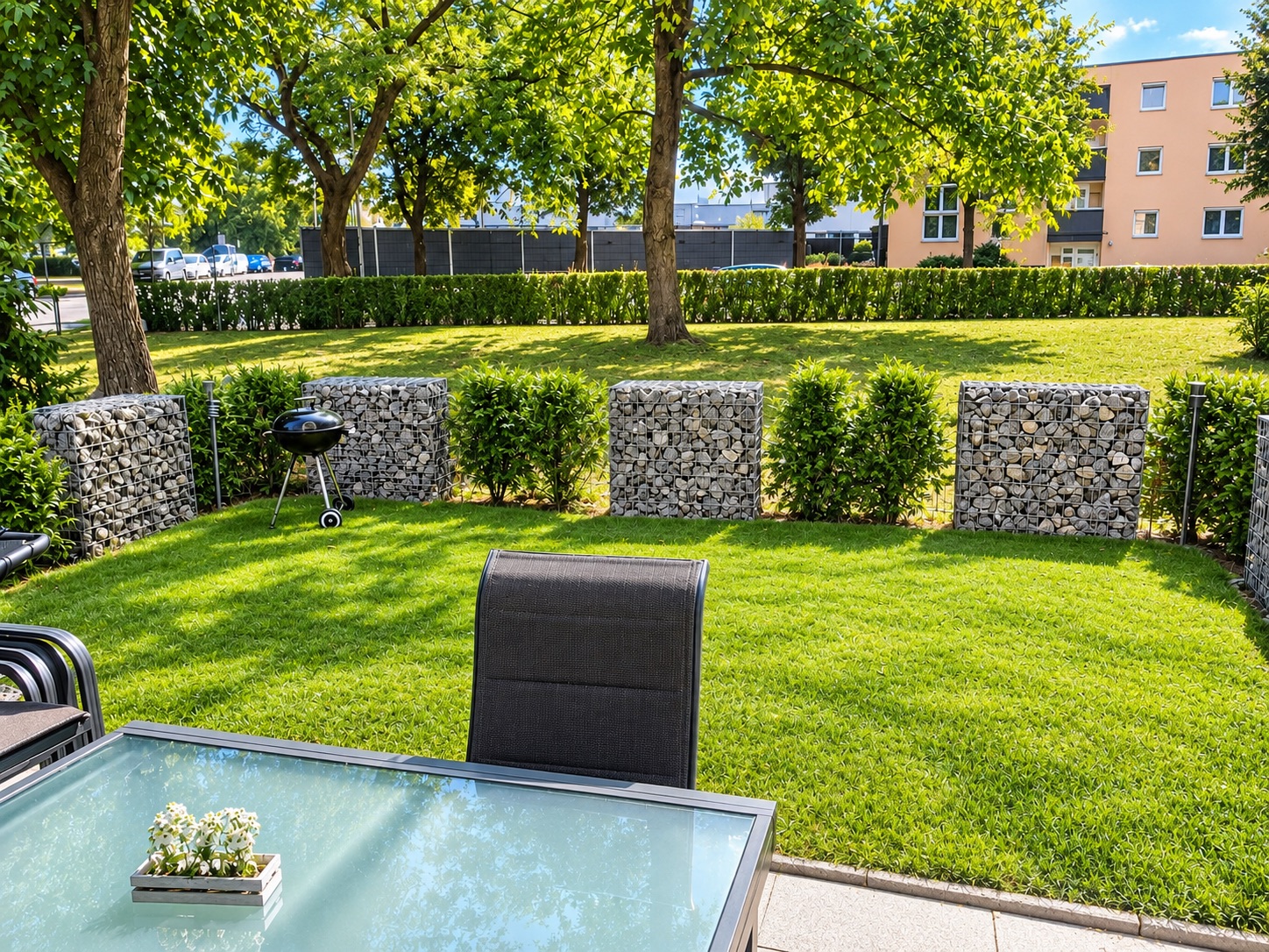Garten mit Rasen, Steinkörben, Grill und Glastisch mit Stuhl im Vordergrund.