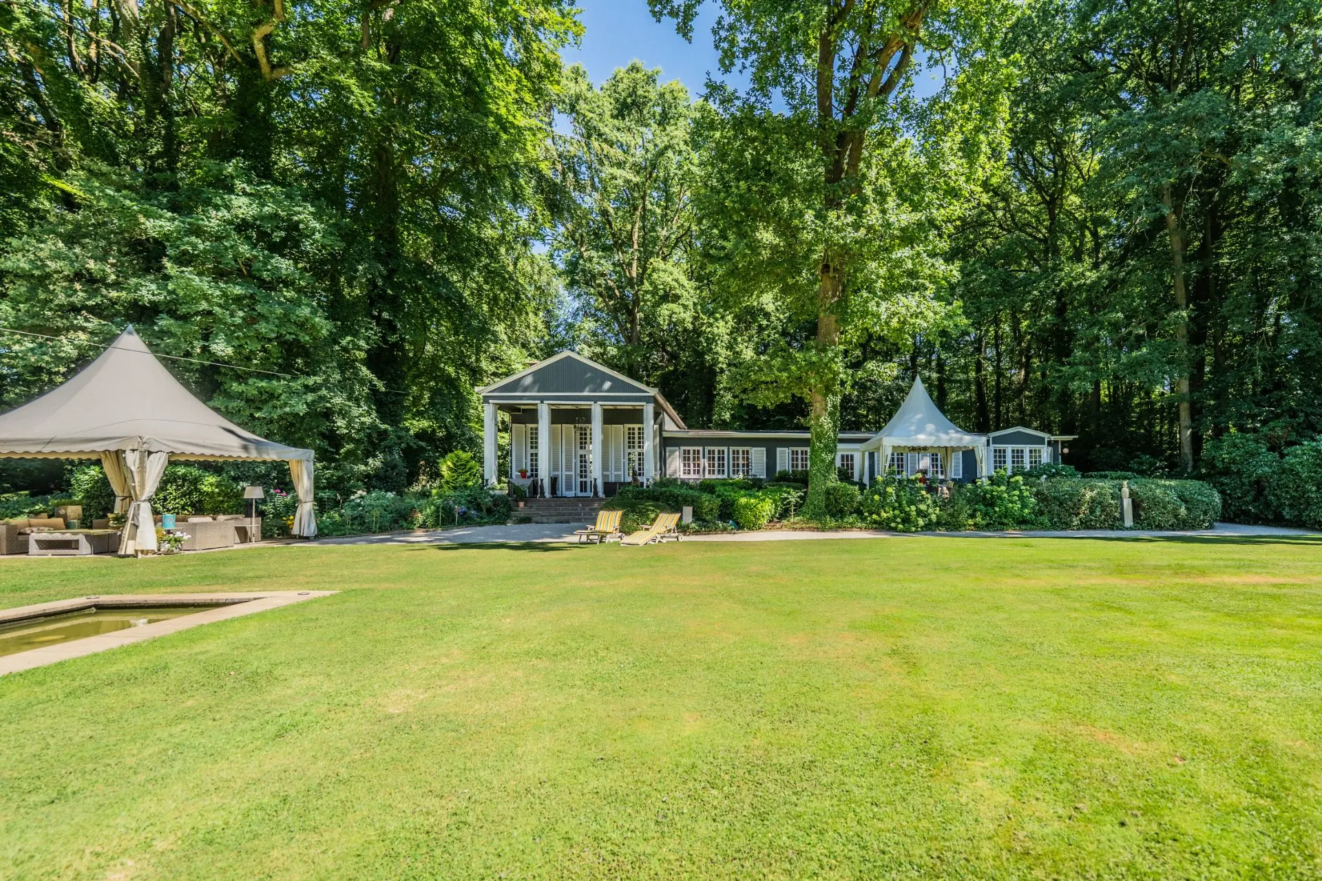 Großes Haus mit Garten, Liegestühlen und zwei Pavillons vor vielen Bäumen.