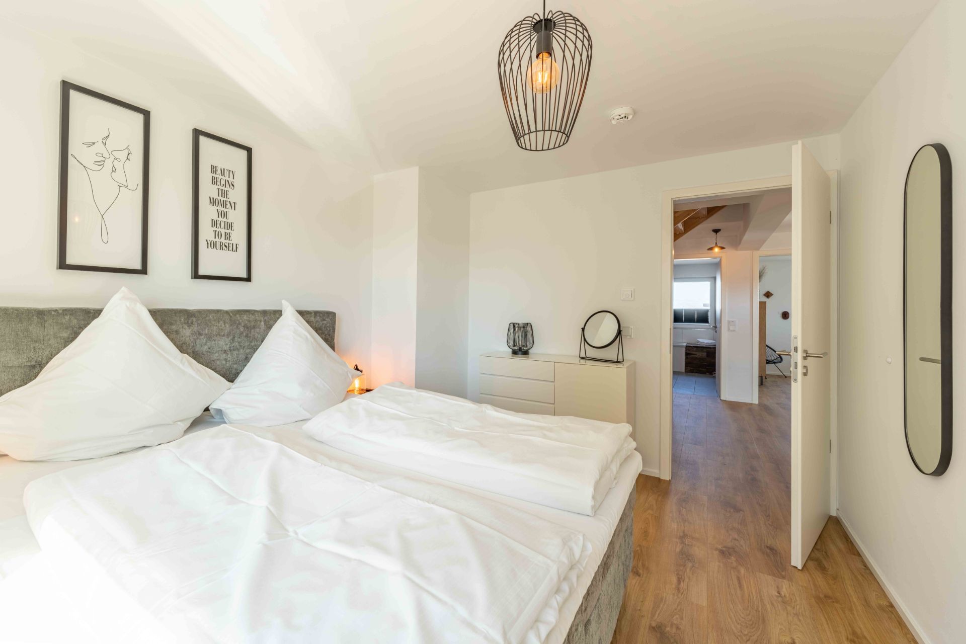 Schlafzimmer mit Doppelbett, zwei Bildern an der Wand und offener Tür zu einem Flur.