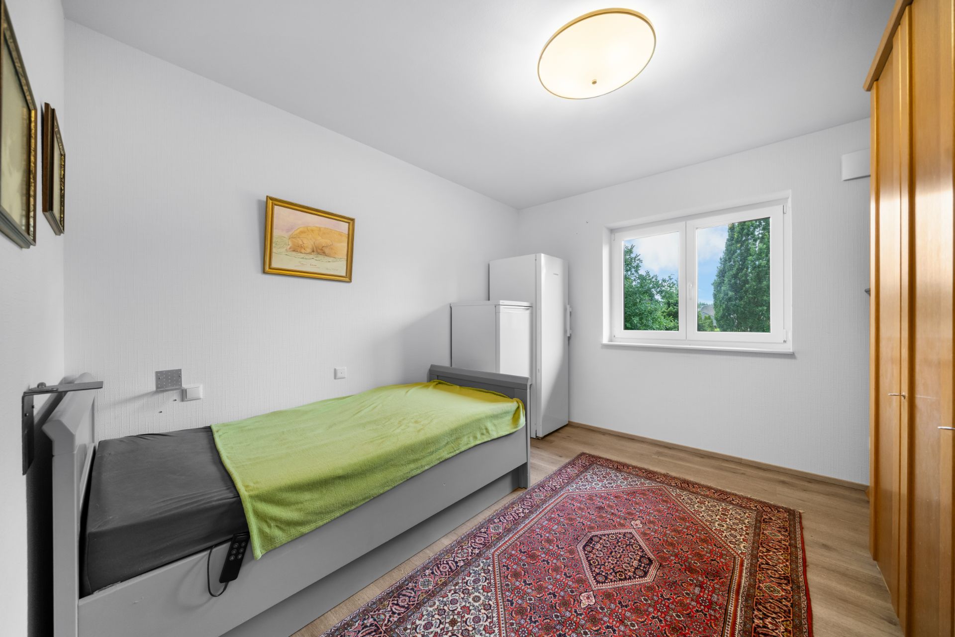 Helles Schlafzimmer mit Einzelbett, Teppich, Kleiderschrank und Fenster mit Blick ins Grüne.