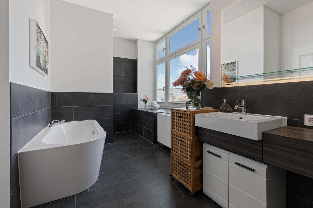 Modernes Badezimmer mit Eckbadewanne, großem Fenster, Waschbecken und dunklen Fliesen.