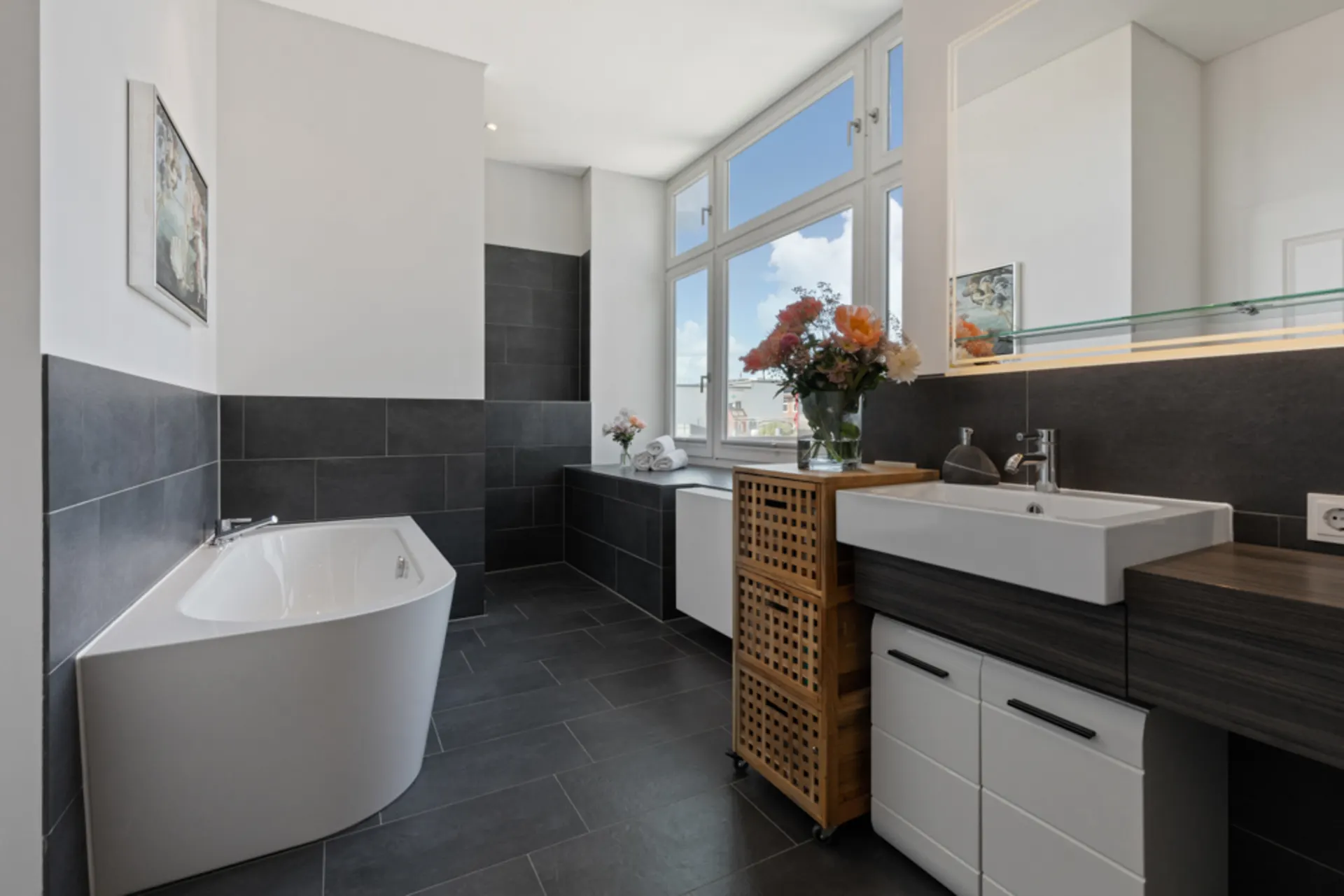 Modernes Badezimmer mit Eckbadewanne, großem Fenster, Waschbecken und dunklen Fliesen.