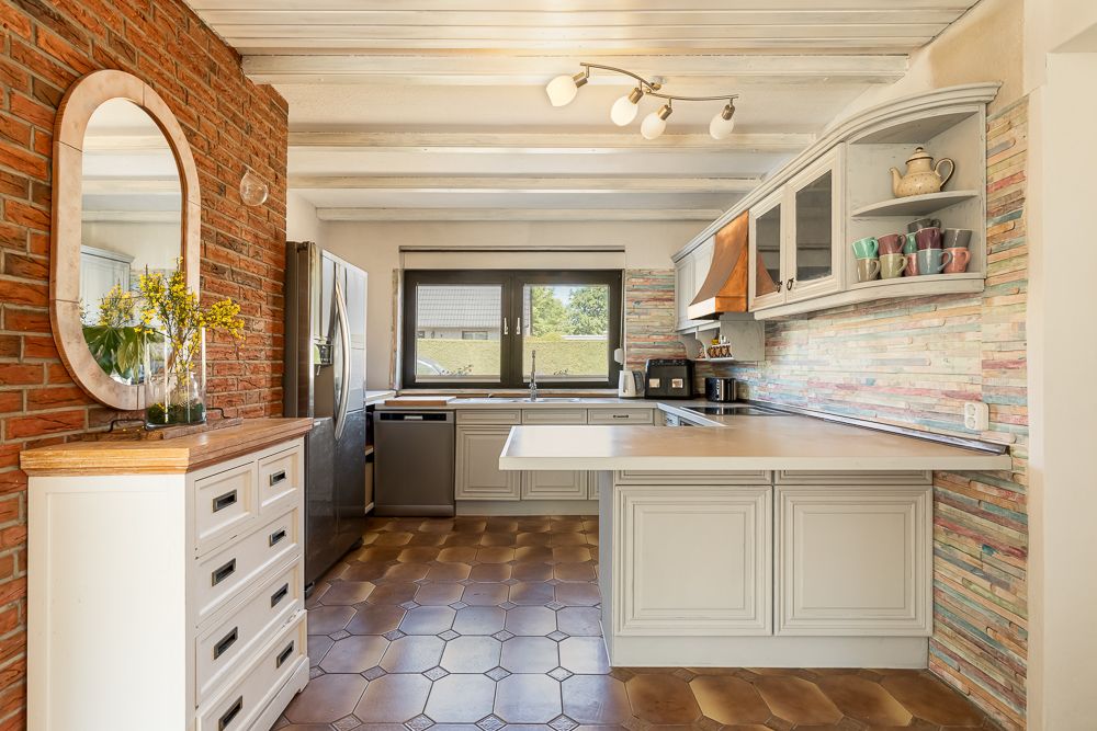 Helle Küche mit weißen Schränken, gemusterter Fliesenwand und Fenstern, Holzdecke und Backsteinwand.